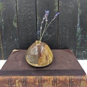 May include: A small, round, brown and white ceramic vase with a single stem of lavender flowers. The vase is sitting on top of a brown leather-bound book.