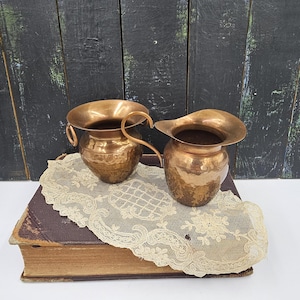 May include: Two small hammered copper pitchers with a handle on one and a flared lip on the other. The pitchers are sitting on a lace doily on top of a brown book.