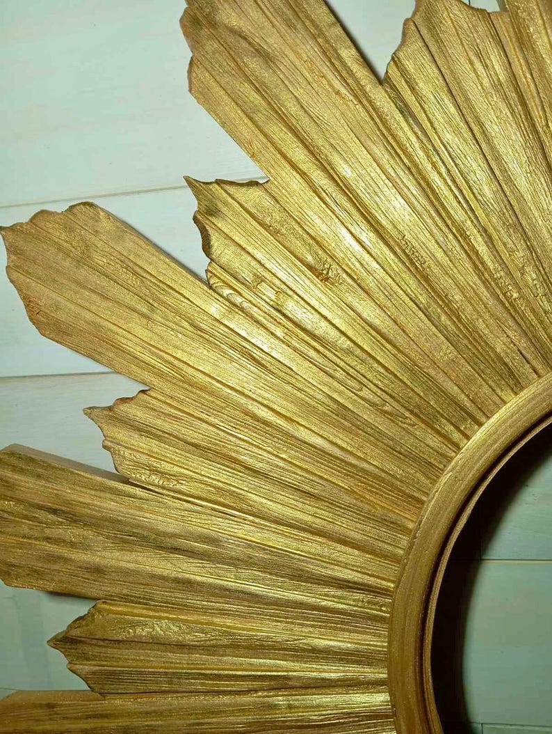 May include: A gold-colored sunburst mirror frame featuring a textured, radiating design with pointed, leaf-like rays extending outward from a central circular opening. The surface has a metallic finish with visible wood-grain-like striations.
