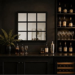 May include: A square wall mirror with a black grid frame featuring nine individual panes. The mirror is mounted on a dark wall above a counter, reflecting a room interior. The frame is composed of thin black lines creating a window-pane design.