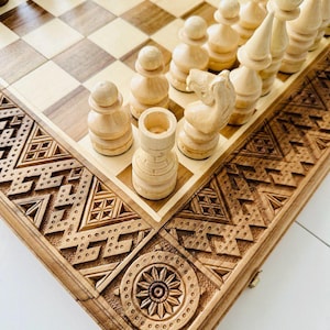 May include: A wooden chessboard with intricate carvings on the edges. The board is made of light and dark wood and has a traditional chessboard pattern. White chess pieces are arranged on the board.