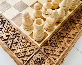 May include: A wooden chessboard with intricate carvings on the edges. The board is made of light and dark wood and has a traditional chessboard pattern. White chess pieces are arranged on the board.