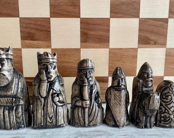 May include: A set of six intricately carved chess pieces in a weathered, brown finish. Each piece depicts a stylized human figure with detailed facial features and regal crowns. The chess pieces are displayed in front of a checkered chessboard.
