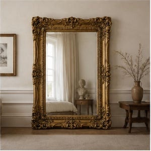 May include: A large rectangular floor mirror featuring an ornate, heavy gold-toned frame with intricate carved floral and scrollwork detailing. The reflective glass surface displays a clear view of a room interior, including a window and furniture.