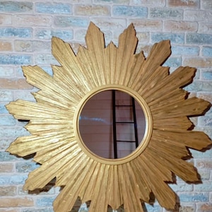 May include: A circular wall mirror featuring a gold-toned sunburst frame with radiating pointed rays. The central glass is round and smooth, surrounded by a textured, gilded wooden structure that extends outward in a star-like pattern.