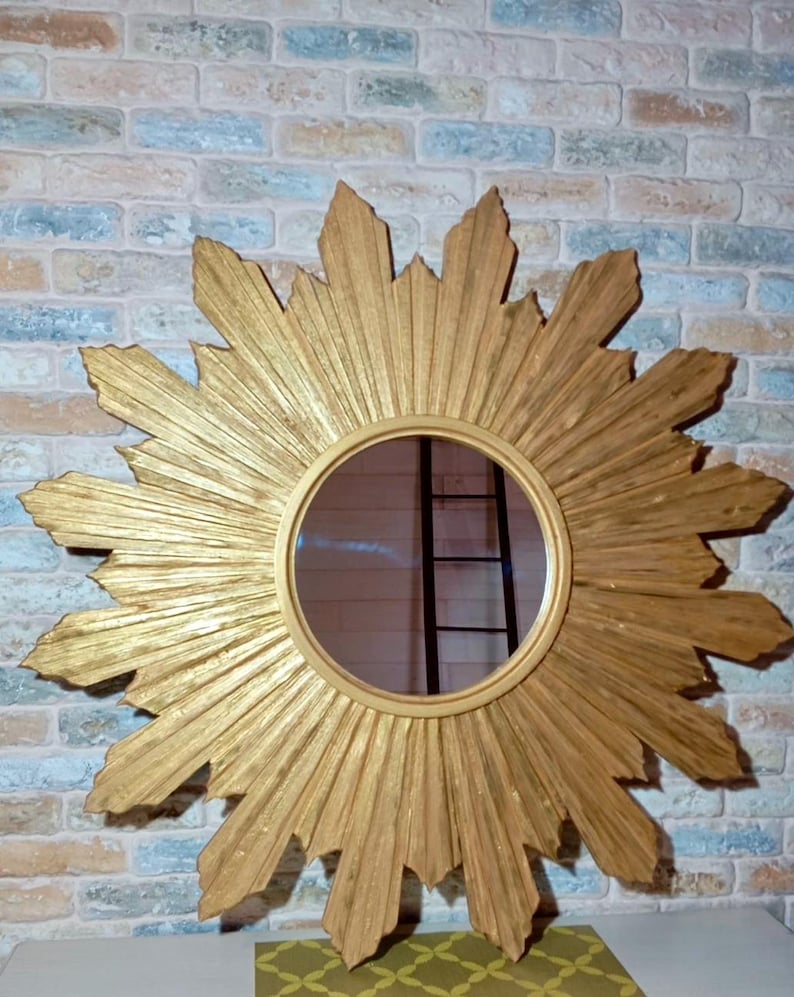 May include: A circular wall mirror featuring a gold-toned sunburst frame with radiating pointed rays. The central glass is round and smooth, surrounded by a textured, gilded wooden structure that extends outward in a star-like pattern.