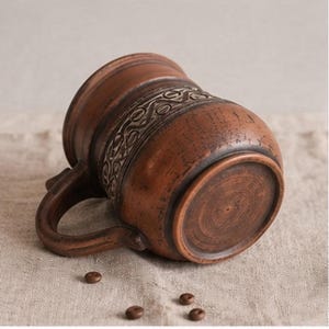 May include: A brown clay mug with a rounded body and a curved handle, featuring a textured band of intricate, repeating geometric patterns around the center. The surface has a rustic, earthy finish with visible variations in the clay color.