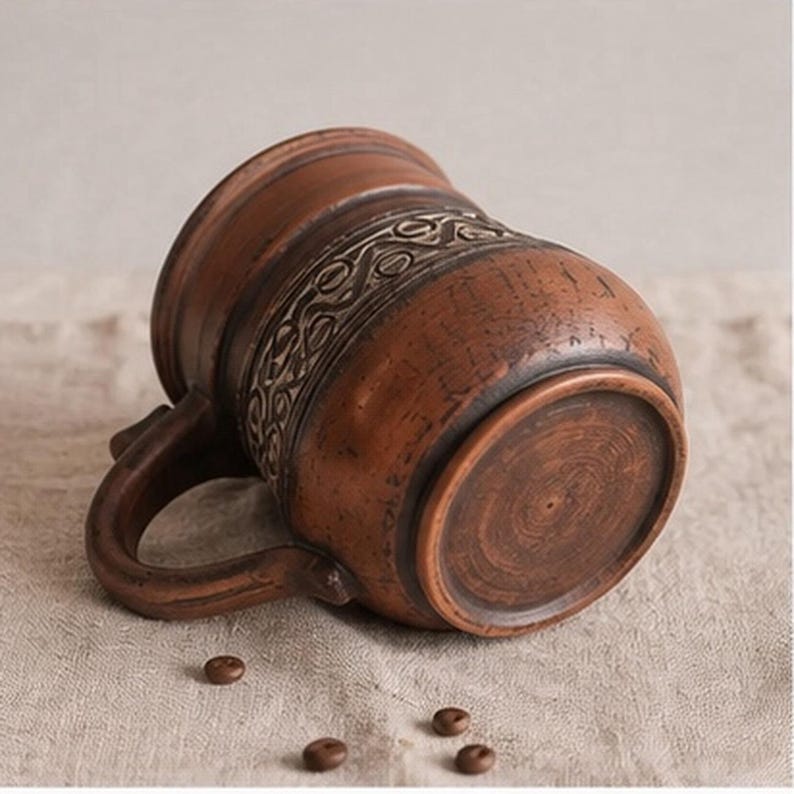 May include: A brown clay mug with a rounded body and a curved handle, featuring a textured band of intricate, repeating geometric patterns around the center. The surface has a rustic, earthy finish with visible variations in the clay color.