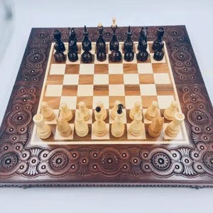 Könnte beinhalten: Ein Schachbrett aus Holz mit einem dunkelbraunen, kunstvoll geschnitzten Rahmen und einer hellen Holzspielfläche. Die Schachfiguren sind in ihren Startpositionen aufgestellt, wobei die weißen Figuren unten und die schwarzen Figuren oben stehen.