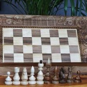 Könnte beinhalten: Ein Schachspiel aus Holz mit aufwendig geschnitzten Details auf der Schachtel. Die Schachfiguren sind aus Holz und auf dem Brett angeordnet. Das Brett besteht aus hellen und dunklen Holzquadraten.