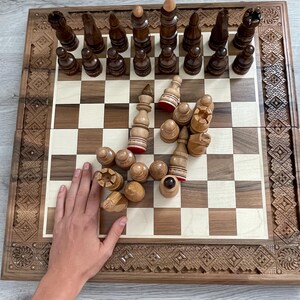 Könnte beinhalten: Ein Schachspiel aus Holz mit aufwendig geschnitzten Details auf dem Brett und den Figuren. Die Schachfiguren sind auf einem hell- und dunkelkarierten Brett angeordnet, wobei sich einige Figuren in der Mitte des Bretts befinden. Das Brett ist aus dunklem Holz mit einem hellen Holzrand.