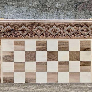 Könnte beinhalten: Ein Schachbrett aus Holz mit einem komplizierten geschnitzten Design am Rand. Das Brett besteht aus hellem und dunklem Holz und bildet ein klassisches Schachbrettmuster.
