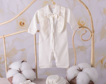 May include: A white baby outfit with a tuxedo-style jacket, a white hat, and two white booties. The outfit is displayed on a white bed with a white cotton boll on each side.