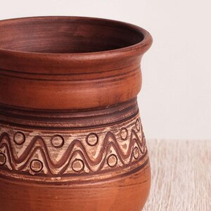 May include: A terracotta-colored clay vessel with a wide, flared rim and a cylindrical body. The exterior features a horizontal band decorated with a repeating pattern of incised zigzag lines and small circles, highlighted with a light-colored wash.