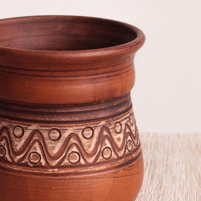 May include: A terracotta-colored clay vessel with a wide, flared rim and a cylindrical body. The exterior features a horizontal band decorated with a repeating pattern of incised zigzag lines and small circles, highlighted with a light-colored wash.