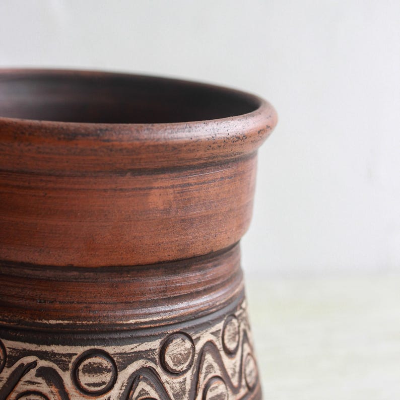 May include: A cylindrical clay vessel with a wide, rounded rim and a smooth, reddish-brown upper section. The lower portion features a dark brown base decorated with a repeating pattern of incised circles and wavy lines in a light beige color.