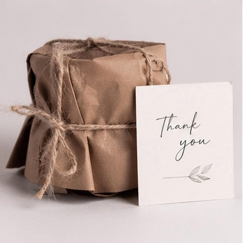 May include: A cylindrical object wrapped in brown kraft paper and secured with thin jute twine, accompanied by a square white card featuring the black printed text Thank you and a small line drawing of a leafy branch at the bottom.