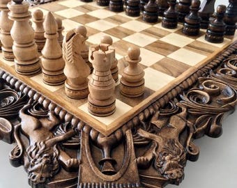 May include: A wooden chess set featuring a board with a light and dark square pattern. The board frame is decorated with intricate carved lion figures and geometric motifs. The set includes light and dark wooden chess pieces arranged on top.