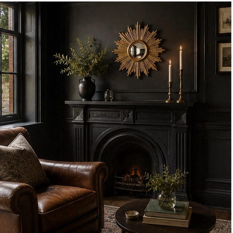 May include: A gold sunburst mirror hangs above a black fireplace mantel. The mantel holds a black vase with greenery and two brass candlesticks with lit white candles. A brown leather armchair sits to the left, and a round table with books sits below.