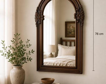 May include: An arched wall mirror with a dark brown wooden frame featuring decorative carved details at the top and sides. The mirror measures 56 cm in width and 76 cm in height, reflecting a bedroom interior with a bed and a lamp.