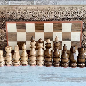 Könnte beinhalten: Ein Satz hölzerner Schachfiguren auf einem Schachbrett aus Holz. Das Schachbrett hat einen geschnitzten Holzrahmen mit aufwendigen Designs. Die Schachfiguren sind aus hellem und dunklem Holz gefertigt und in einer traditionellen Schachstellung angeordnet.