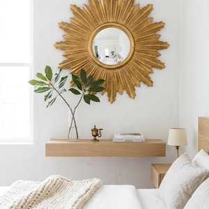 May include: A large gold sunburst mirror with a circular center glass, mounted on a white wall above a light wood floating shelf. The shelf holds a clear glass vase with green leaves, a small brass object, and two stacked books.