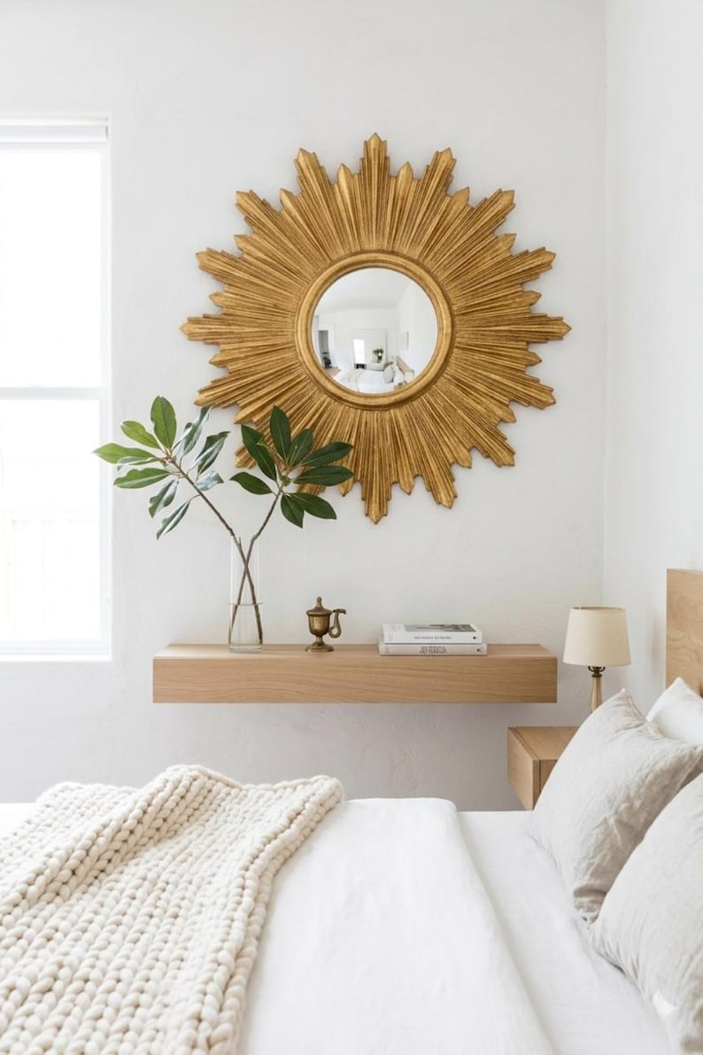 May include: A large gold sunburst mirror with a circular center glass, mounted on a white wall above a light wood floating shelf. The shelf holds a clear glass vase with green leaves, a small brass object, and two stacked books.