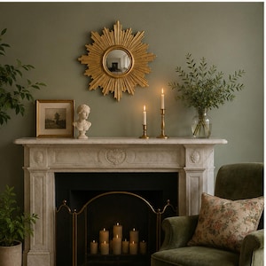 May include: A gold sunburst mirror with a circular center, featuring radiating rays of varying lengths. The mirror is mounted on a wall above a white marble fireplace mantel, which holds two brass candlesticks with lit white taper candles.