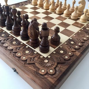 Könnte beinhalten: Ein Schachspiel aus Holz mit aufwendigen Schnitzereien auf dem Brett und den Figuren. Das Brett ist aus dunklem Holz mit einer Einlage aus hellem Holz. Die Figuren sind aus dunklem und hellem Holz. Das Schachspiel ist in geschlossener Position, wobei die Figuren in ihren Startpositionen angeordnet sind.