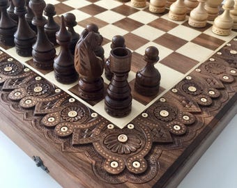 May include: A wooden chess set with intricate carvings on the board and pieces. The board is made of dark wood with a light wood inlay. The pieces are made of dark wood and light wood. The chess set is in a closed position, with the pieces arranged in their starting positions.