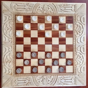 May include: A wooden checkerboard game with a carved frame and white and grey checkers. The board has a light brown and dark brown checkered pattern.