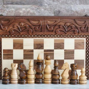 Könnte beinhalten: Ein Schachspiel aus Holz mit aufwendig geschnitzten Figuren und einem Brett aus Holz mit Nussbaum- und Ahornfurnier. Das Schachbrett ist in einer Holzkiste mit einem geschnitzten Design verschlossen.
