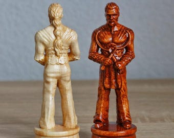May include: Two wooden chess pieces, one light brown and one dark brown, are standing on a wooden surface. The light brown piece is a knight with a long braid and is facing forward. The dark brown piece is a knight with a beard and is holding a weapon. Both pieces are carved in detail.