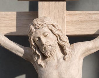 May include: A light-colored wooden crucifix featuring a detailed carving of a figure with long hair and a beard. The figure is positioned on a cross with a horizontal beam above the head that displays the carved letters INRI.