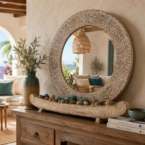 May include: A round wall mirror with a wide, intricately carved wooden frame featuring geometric patterns. Below the mirror sits a long, curved wooden decorative bowl filled with small, round, textured spheres in shades of blue and brown.