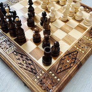 Könnte beinhalten: Ein Schachspiel aus Holz mit aufwendigen Schnitzereien auf dem Brett und den Figuren. Das Brett ist aus hellem und dunklem Holz, die Figuren aus dunklem Holz. Die Figuren sind in der Startposition auf dem Brett angeordnet.