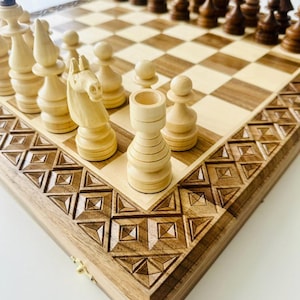 Könnte beinhalten: Ein Schachspiel aus Holz mit hell- und dunkelbraunen Feldern und einem aufwendig geschnitzten Rand. Die Schachfiguren sind hell- und dunkelbraun und auf dem Brett angeordnet. Das Set wird auf einer weißen Oberfläche präsentiert.