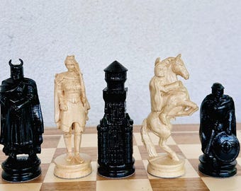 May include: A set of five detailed chess pieces including three black figures and two cream-coloured figures. The pieces depict a horned knight, a standing figure, a tall castle tower, a rider on a rearing horse, and a soldier holding a round shield.