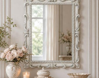 May include: A rectangular wall mirror with an ornate, off-white frame featuring intricate scrollwork and beaded detailing. The mirror reflects a bright room with a window and curtains, while the frame displays a weathered, textured finish.