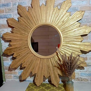 May include: A large circular wall mirror featuring a gold-toned sunburst frame with radiating pointed rays. The mirror has a smooth, round central glass surface. A small glass vase containing dried brown grass sits in front of the mirror.