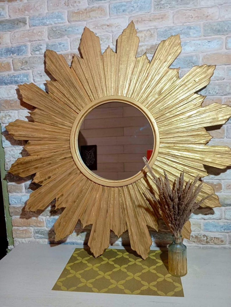 May include: A large circular wall mirror featuring a gold-toned sunburst frame with radiating pointed rays. The mirror has a smooth, round central glass surface. A small glass vase containing dried brown grass sits in front of the mirror.