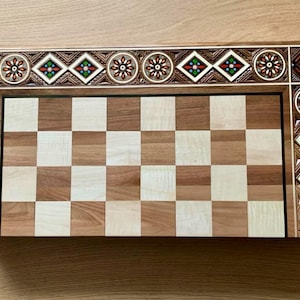 Könnte beinhalten: Ein Schachbrett aus Holz mit einem dekorativen, eingelegten Rand. Das Brett hat ein traditionelles Schachbrettmuster mit abwechselnd hellen und dunklen Feldern.