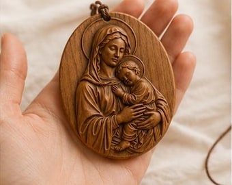 May include: An oval-shaped wooden pendant featuring a detailed relief carving of a figure holding a child, both with circular halos. The brown wood piece has a smooth finish and is attached to a thin, dark brown cord for wearing as a necklace.
