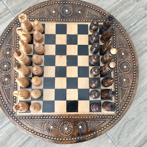 Könnte beinhalten: Ein Schachspiel aus Holz mit aufwendigen Schnitzereien auf dem runden Brett. Die Schachfiguren sind in ihren Startpositionen auf dem Brett angeordnet, wobei die weißen Figuren links und die schwarzen Figuren rechts stehen.