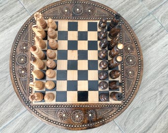 May include: A wooden chessboard set with intricate carvings on the circular board. The chess pieces are arranged in their starting positions on the board, with the white pieces on the left and the black pieces on the right.