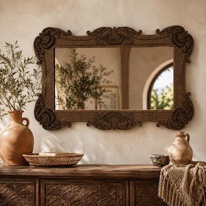 Pode incluir: Um espelho de parede retangular com uma moldura de madeira esculpida em castanho escuro, decorada com volutas ornamentadas e motivos em forma de concha nos cantos e nas bordas centrais. A superfície de vidro reflete o interior.