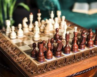 May include: A wooden chessboard with intricate carvings around the edge. The board is open and shows a game in progress with dark brown and cream coloured pieces.
