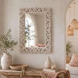 Puede incluir: Un espejo de pared rectangular con un marco de madera clara desgastada que presenta intrincados patrones tallados de flores y volutas. La superficie del espejo refleja un espacio interior luminoso con una ventana y elementos decorativos neutros.