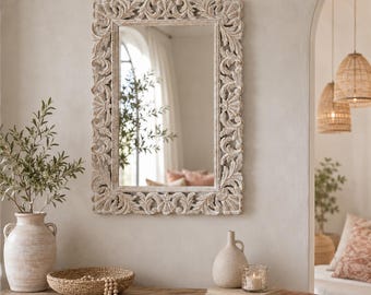 May include: A rectangular wall mirror featuring a light-colored, weathered wood frame with intricate, carved floral and scrollwork patterns. The mirror surface reflects a bright interior space with a window and soft, neutral-toned decor elements.