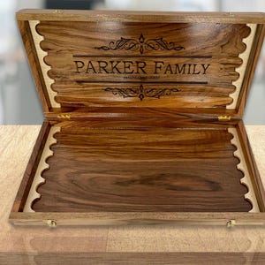 Könnte beinhalten: Eine Holzkiste für Spiele mit einer hellbraunen Oberfläche und einem gewellten Rand. Der Deckel ist geöffnet und das Innere ist mit hellem Holz ausgekleidet. Der Deckel ist mit dem Text "PARKER FAMILY" und einem dekorativen Muster graviert.